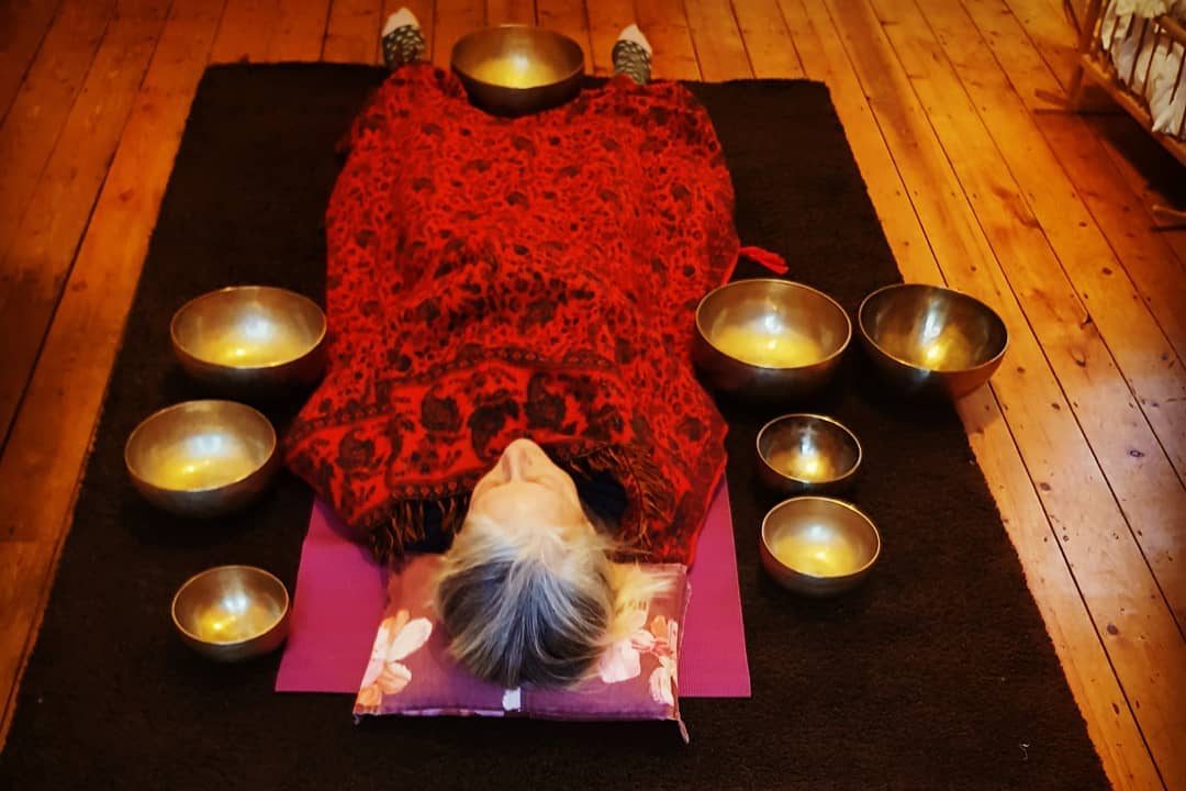 A person lying wrapped in a red blanket surrounded by a circle of Tibetan singing bowls